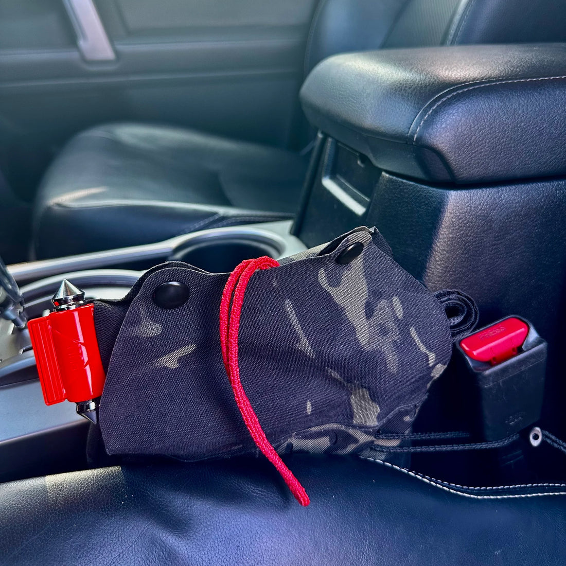 A black and gray camouflage car accessory pouch with a red drawstring, resting on a black leather car seat near the center console.