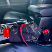 A black and gray camouflage car accessory pouch with a red drawstring, resting on a black leather car seat near the center console.